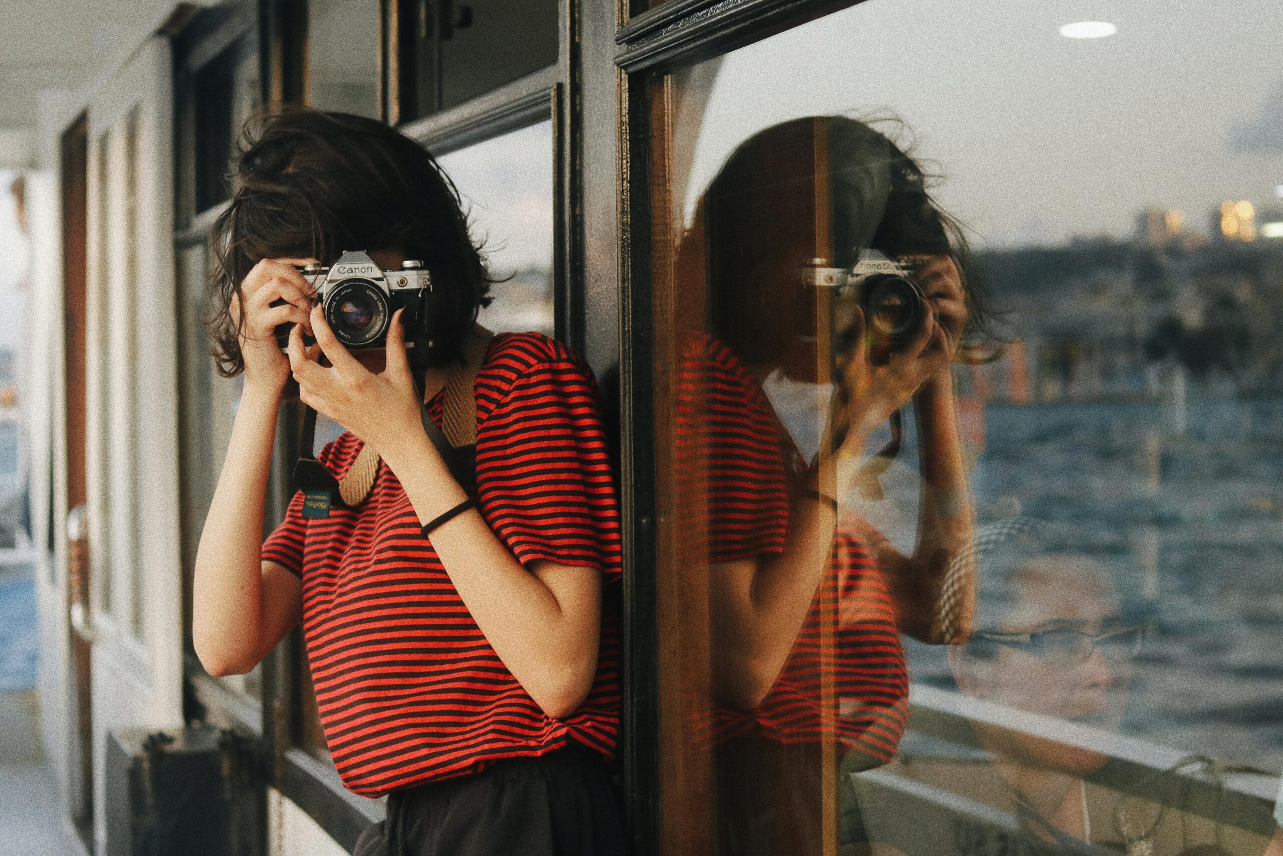 Woman photographing through a ferry window, reflected in the glass, holding a film camera