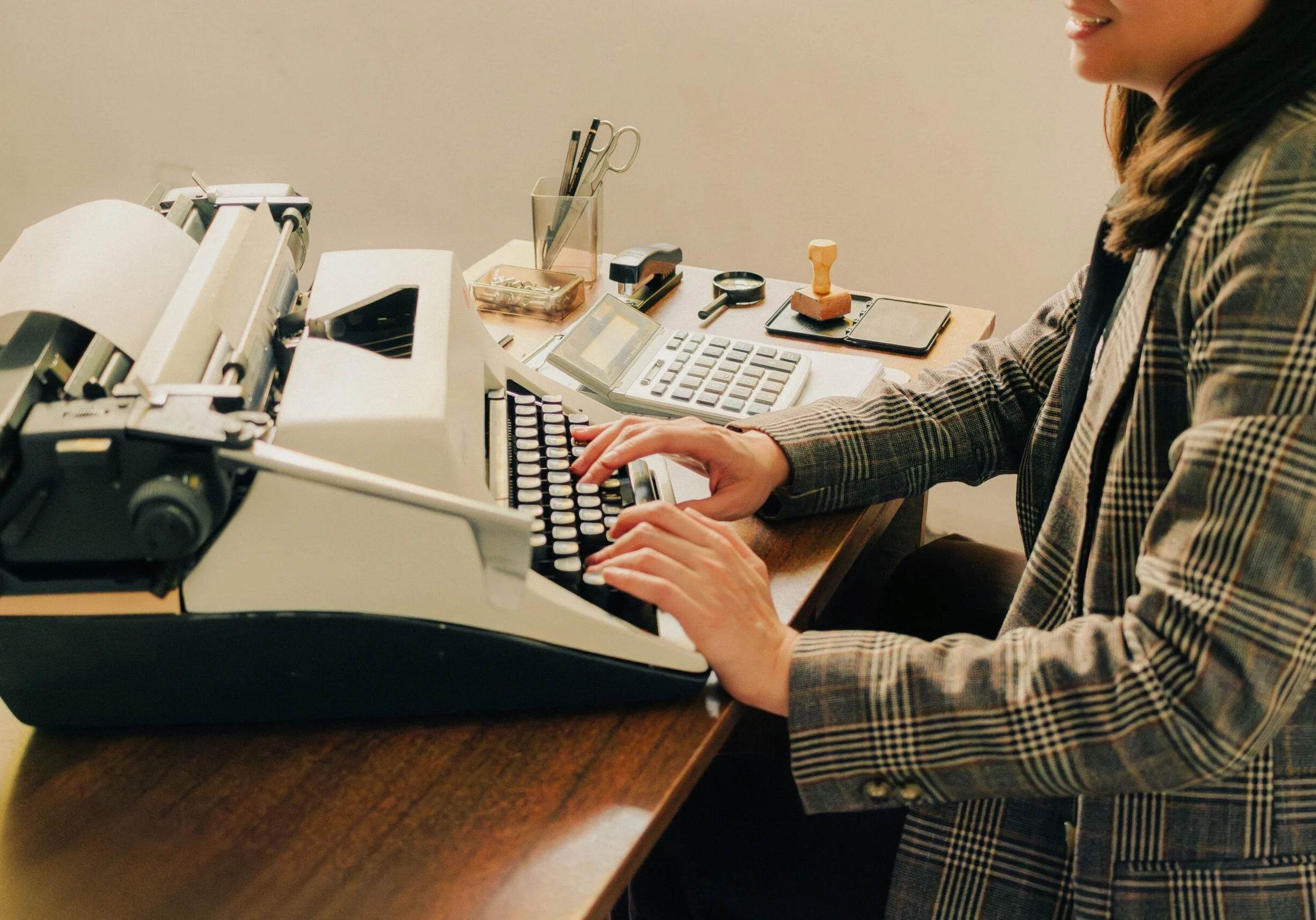 Woman in a plaid blazer typing on a vintage typewriter at a wooden desk
