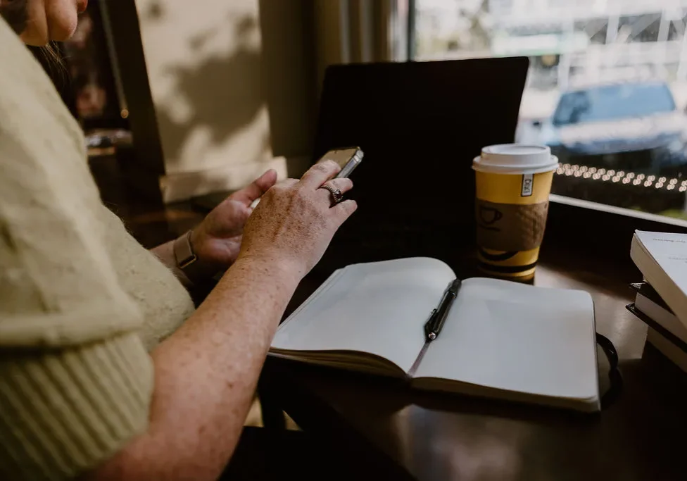 Woman entrepreneur reviewing marketing strategy on her phone at a cafe in Saint John NB, with an open notebook and laptop